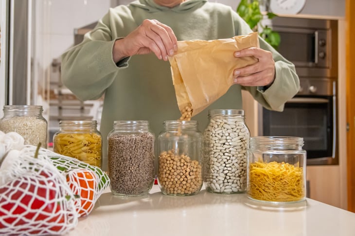 Person filling glass jars with non-perishable bulk foods, like beans, rice, and pasta.