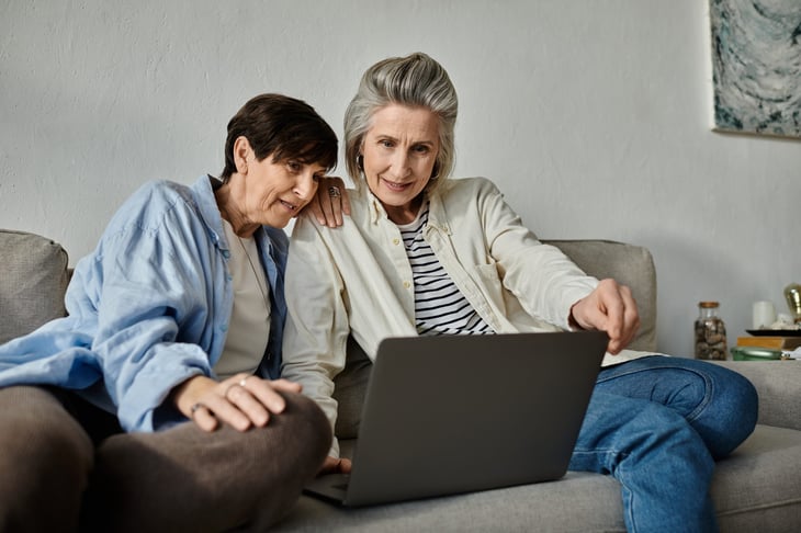 Two older women look at a laptop while sitting on a couch.