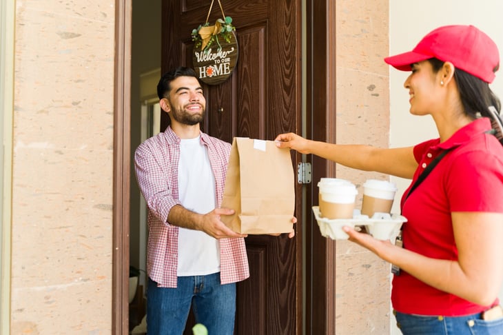 Delivery woman is handing a bag of food and coffee to a customer at his front door
