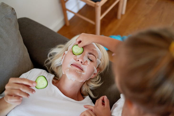 Little girl helps her mom put on a sheet mask and cucumbers on her eyes.