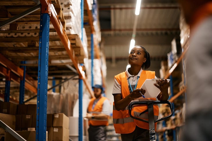 Woman working and looking at a clipboard in a warehouse with a coworker in the background