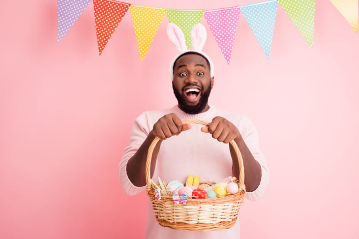 A smiling man wearing bunny ears and holding an Easter basket full of candy, eggs, and cookies.
