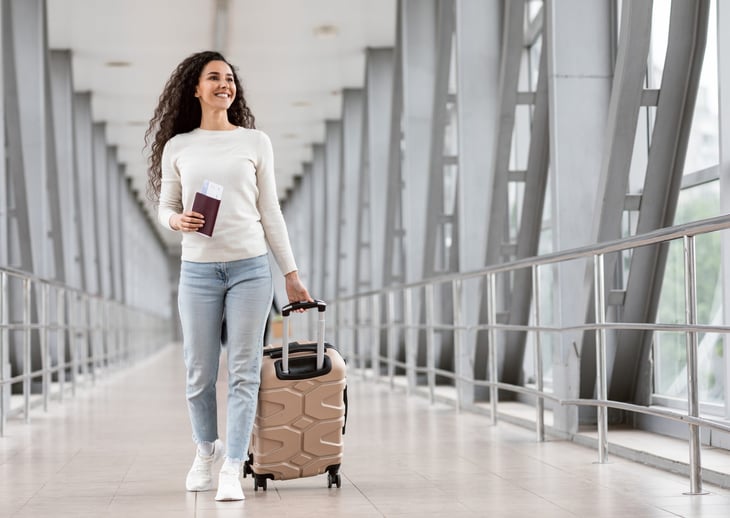 Smiling woman walking through an airport terminal with luggage, passport, and ticket.