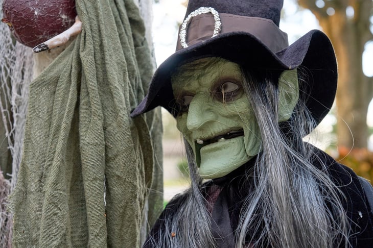 Green witch prop with black hat and silver buckle, long gray hair, and cracked green skin. Spooky Halloween yard decoration photographed outdoors in daylight.