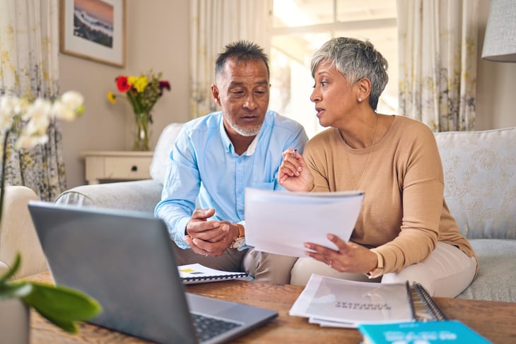 Senior couple on a couch at home with a laptop, bills and documents. Going over finances.