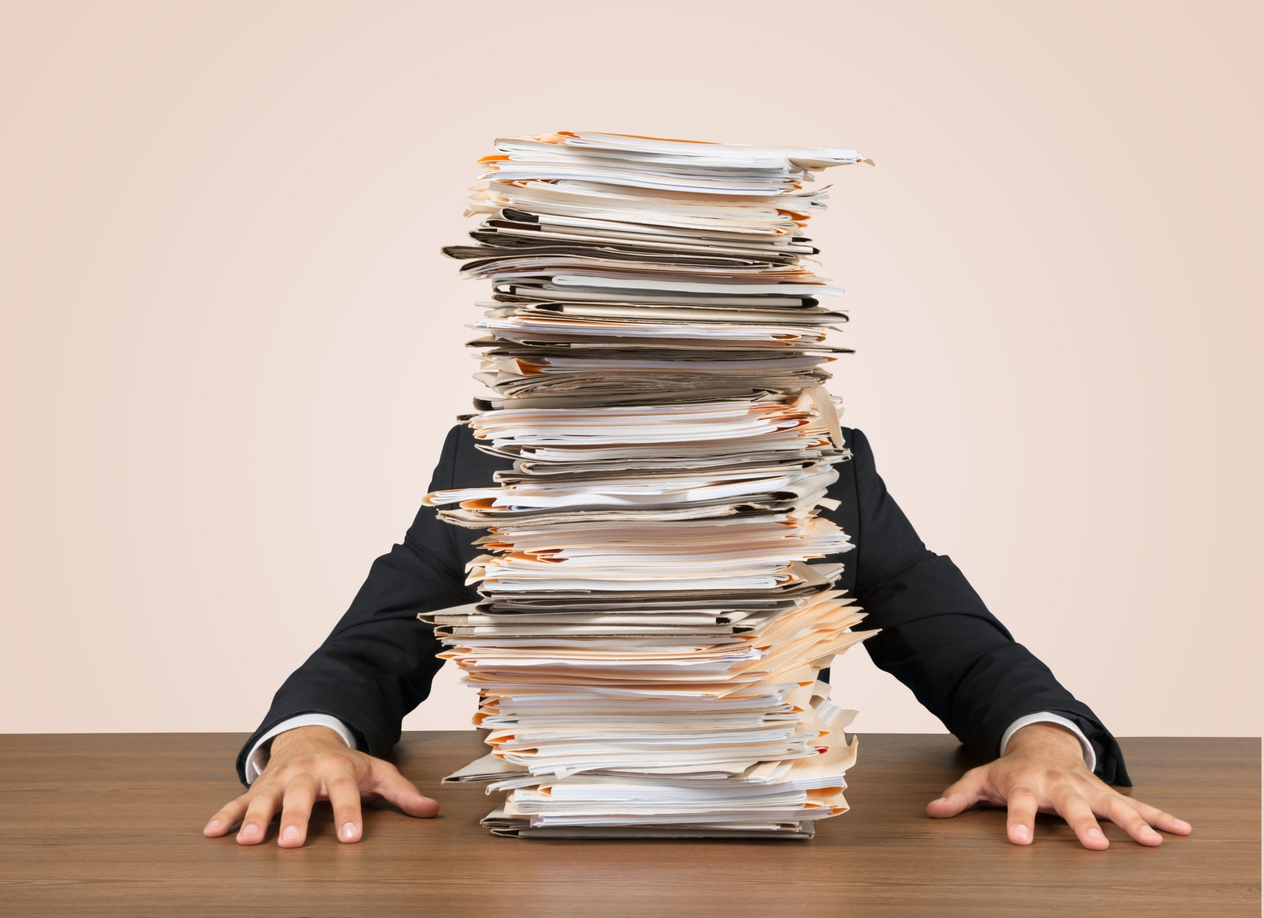  A person in a suit is sitting at a desk with a large stack of documents and paperwork piled high in front of them, symbolizing the search query 'Documents and paperwork for insurance claim'.